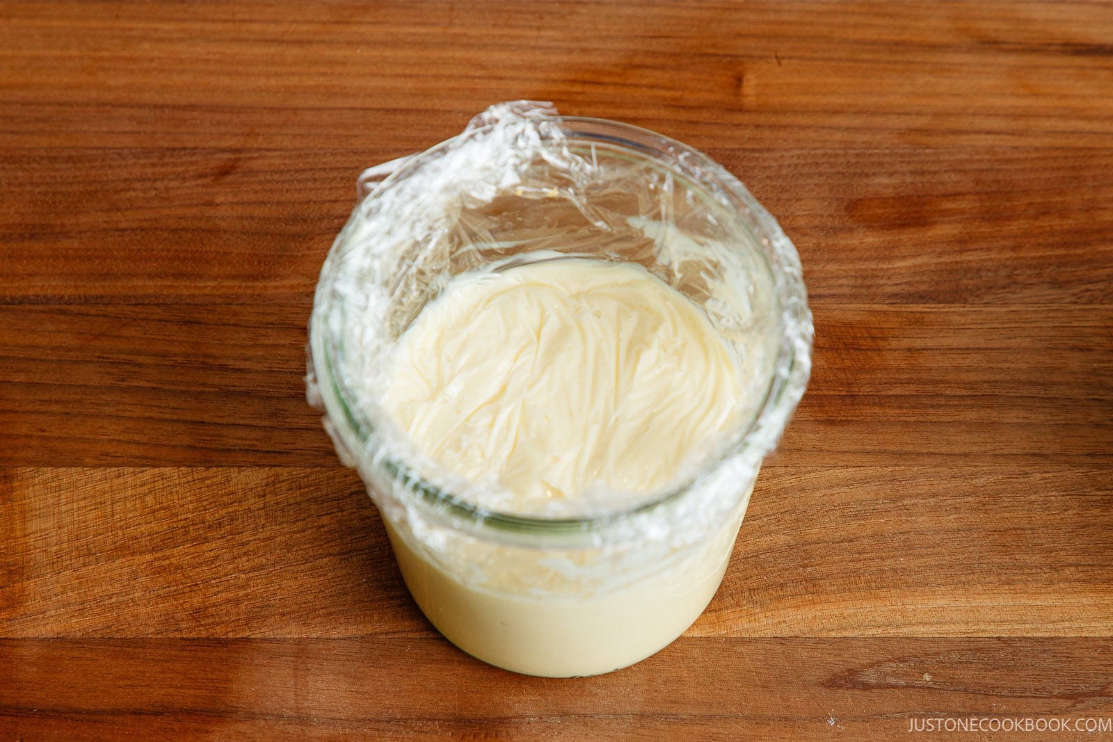 A glass bowl of creamy custard, enhanced with a touch of Japanese mayonnaise, sits covered with plastic wrap on a wooden surface.