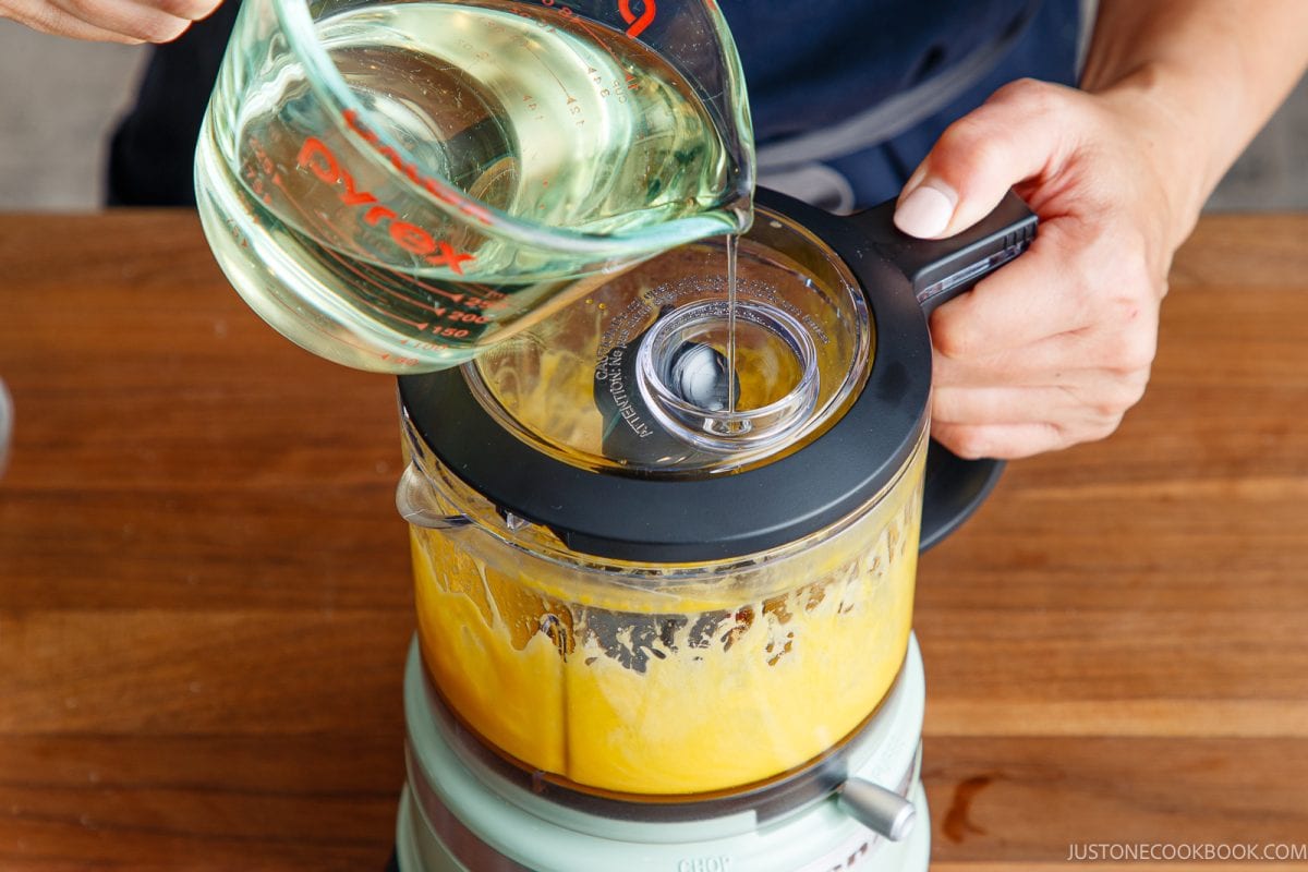 A person pours oil from a measuring cup into a running food processor filled with japanese mayonnaise mixture on a wooden table.