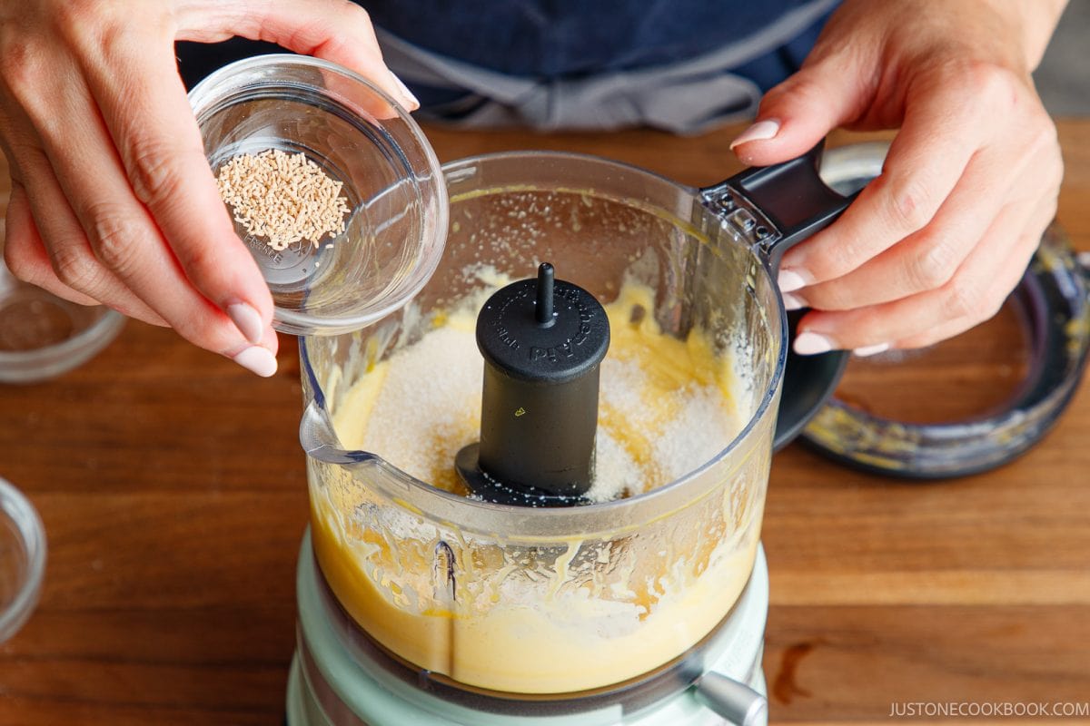 A person pours dashi powder from a small bowl into a food processor containing japanese mayonnaise with their other hand holding the processor lid open.