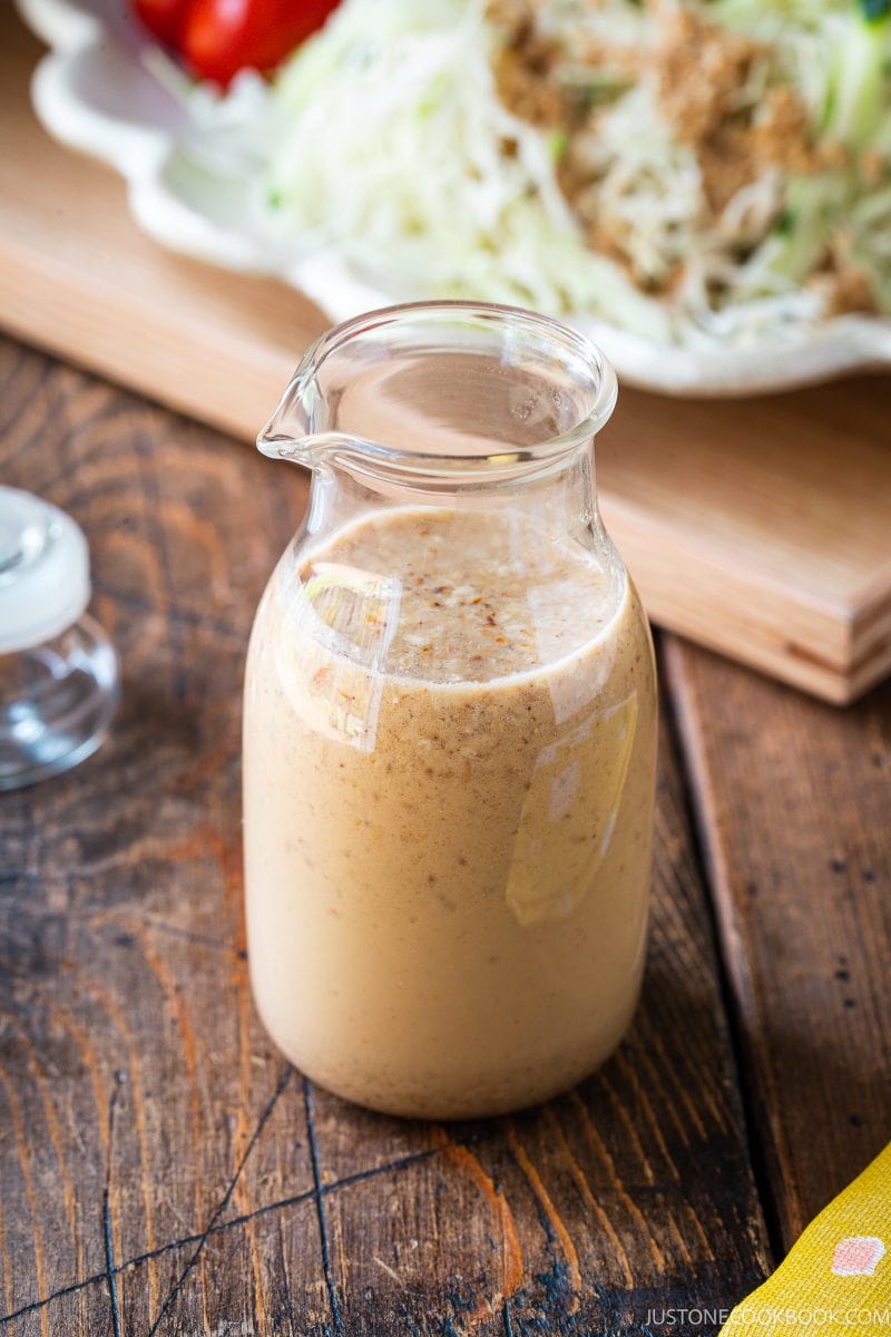 A small glass pitcher filled with creamy, light brown salad dressing sits on a rustic wooden table, with a plate of shredded vegetables blurred in the background.