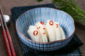 A bowl of thinly sliced pickled daikon garnished with red chili rings, served in a blue and white striped bowl on a dark patterned cloth, with chopsticks beside it.