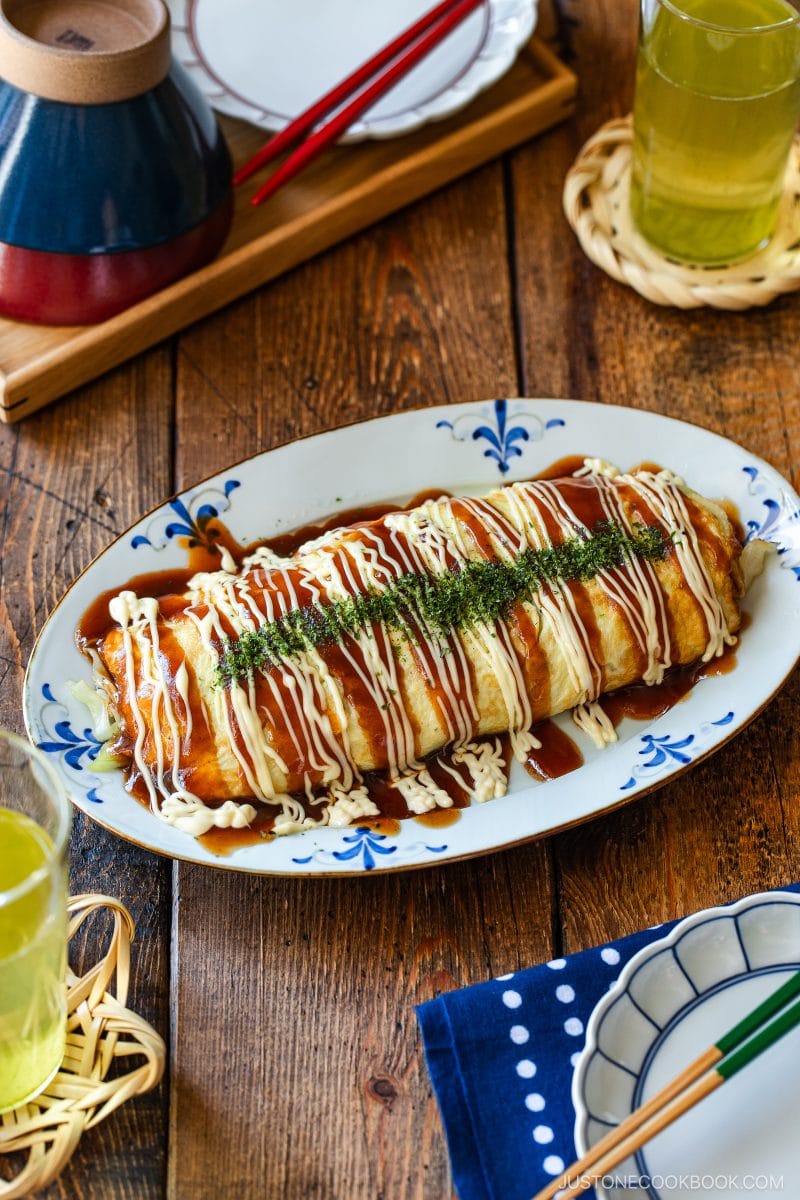 A Japanese tonpeiyaki (pork and cabbage omelette), topped with mayonnaise, okonomiyaki sauce, and green seaweed, served on a white and blue platter with drinks and side dishes on a wooden table.