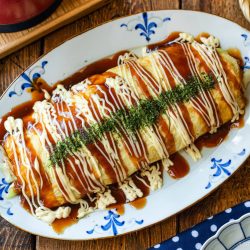 A Japanese tonpeiyaki omelette topped with savory sauces and creamy mayonnaise, garnished with green seaweed flakes, served on a decorative oval plate on a wooden table with chopsticks and side dishes nearby.