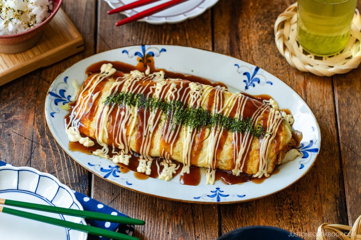 A large Japanese omelette, known as tonpeiyaki (pork and cabbage roll), is topped with zigzag lines of Japanese mayonnaise, okonomi sauce, and dried seaweed flakes, and served on an oval plate on a wooden table.