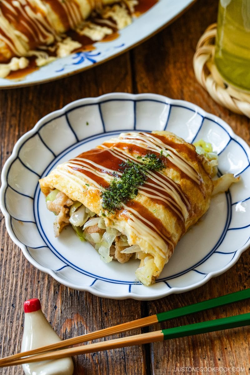 A plate with sliced Japanese tonpeiyaki (pork and cabbage omelette), topped with mayonnaise, okonomi sauce, and green seaweed, revealing a savory filling of cabbage and meat inside with chopsticks and a sauce dispenser nearby on a wooden table.