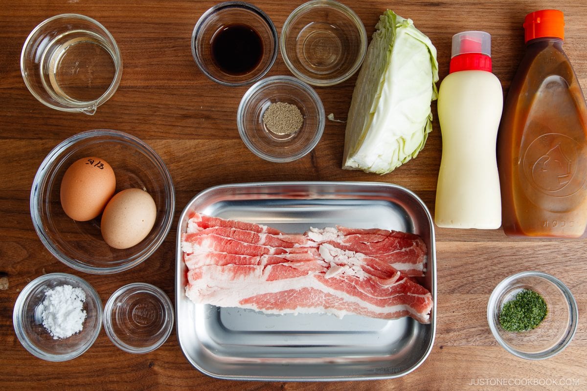 Top-down view of ingredients for Japanese tonpeiyaki, including eggs, pork belly slices, cabbage, sauces, seasonings, oil, and flour, all arranged neatly on a wooden surface.