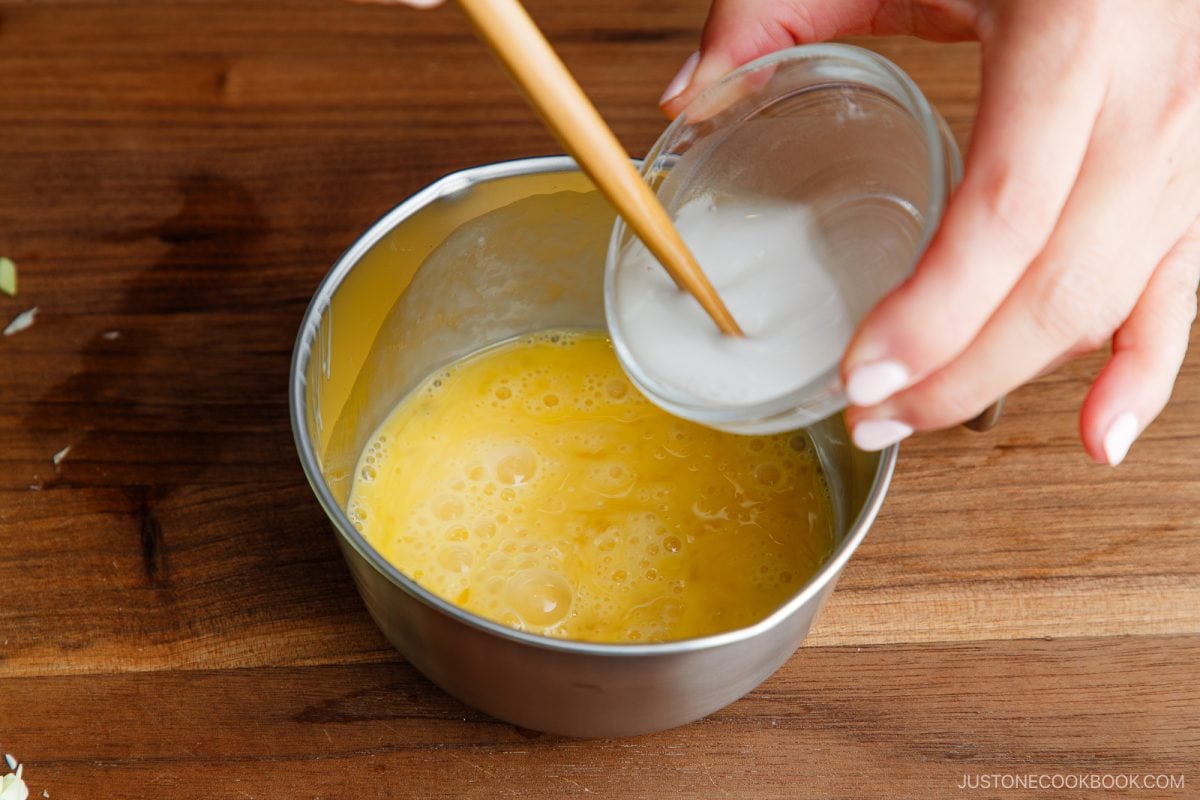 A hand pours a small bowl of white liquid into a bowl of beaten eggs on a wooden surface, using a wooden chopstick to stir&mdash;perfectly preparing the egg mixture for making tonpeiyaki.
