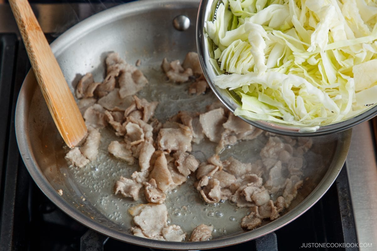 A frying pan with cooked pork slices on a stove, a wooden spatula resting on the side, and sliced cabbage being added from a bowl above the pan&mdash;just like preparing classic tonpeiyaki.