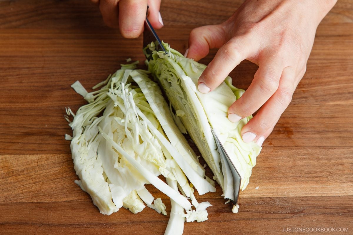 Hands slicing a head of cabbage with a knife on a wooden cutting board, preparing fresh ingredients for tonpeiyaki. Thin cabbage strips are visible beside the hands.
