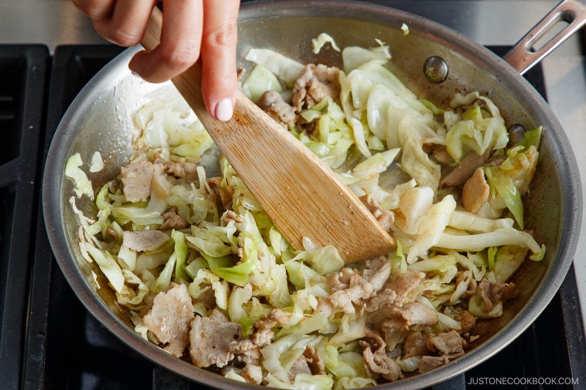A hand stirs sliced cabbage and pieces of meat in a skillet with a wooden spatula on a stovetop, preparing a savory tonpeiyaki.