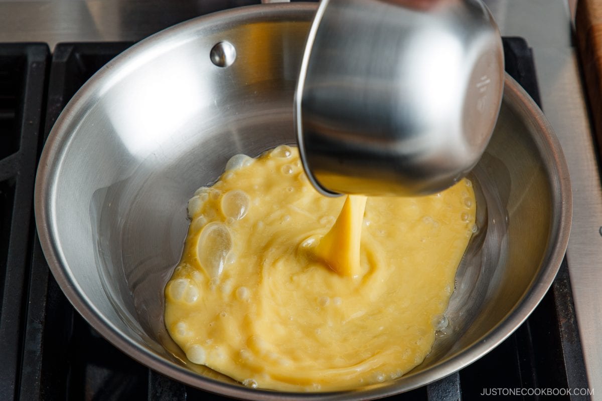Beaten eggs are being poured from a metal bowl into a stainless steel frying pan on a stovetop, ready to be cooked into tonpeiyaki.