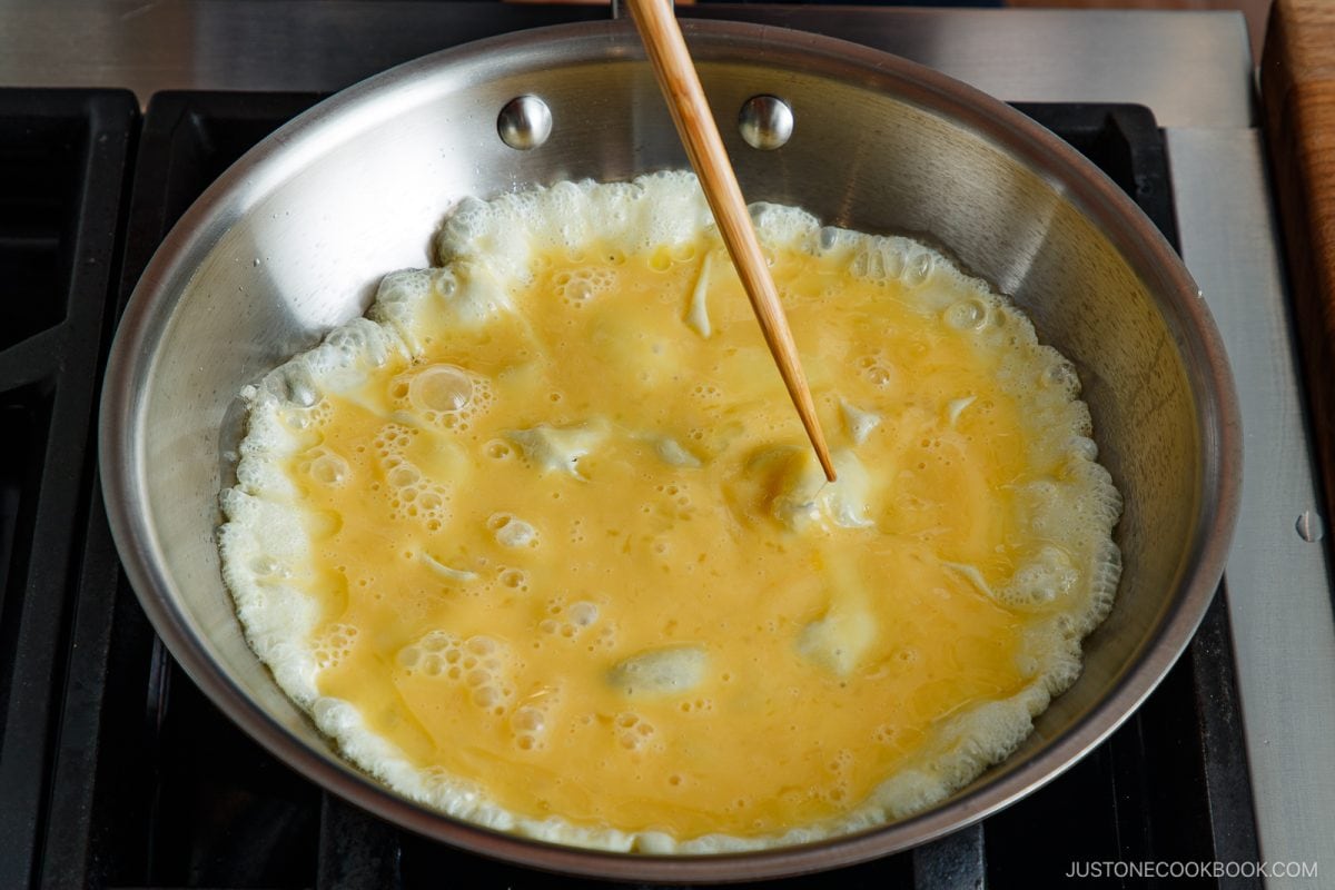 A pair of chopsticks stirs beaten eggs cooking in a large stainless steel pan on a stovetop, preparing the perfect base for a savory tonpeiyaki.