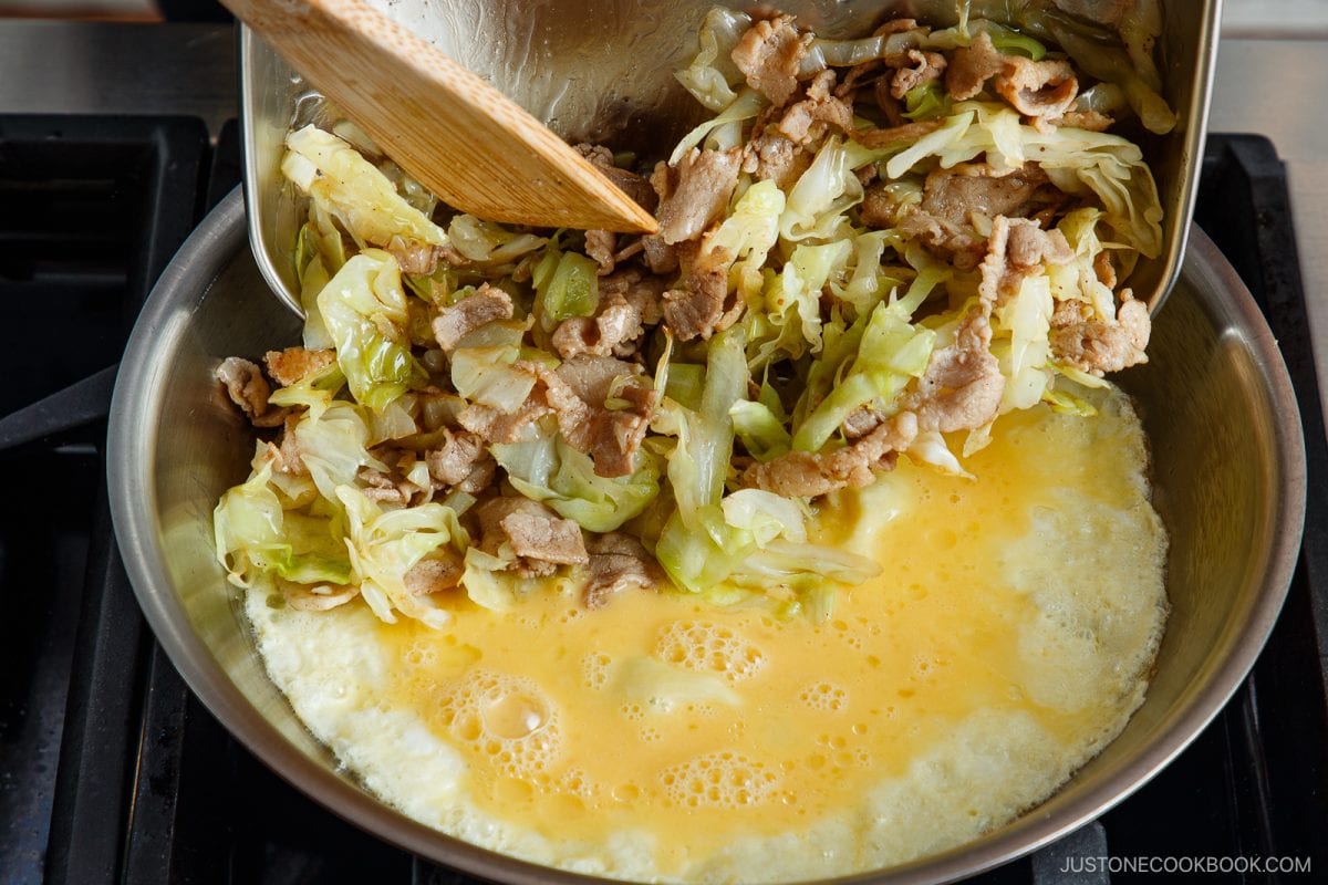 A pan with beaten eggs cooking, reminiscent of making tonpeiyaki, while stir-fried pork and cabbage are added from a bowl using a wooden spatula. The dish is being prepared on a stovetop.