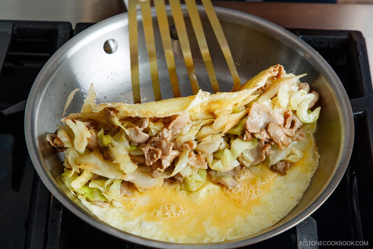 A stainless steel pan on a stove with a tonpeiyaki omelette being folded over stir-fried pork and cabbage using a slotted spatula.