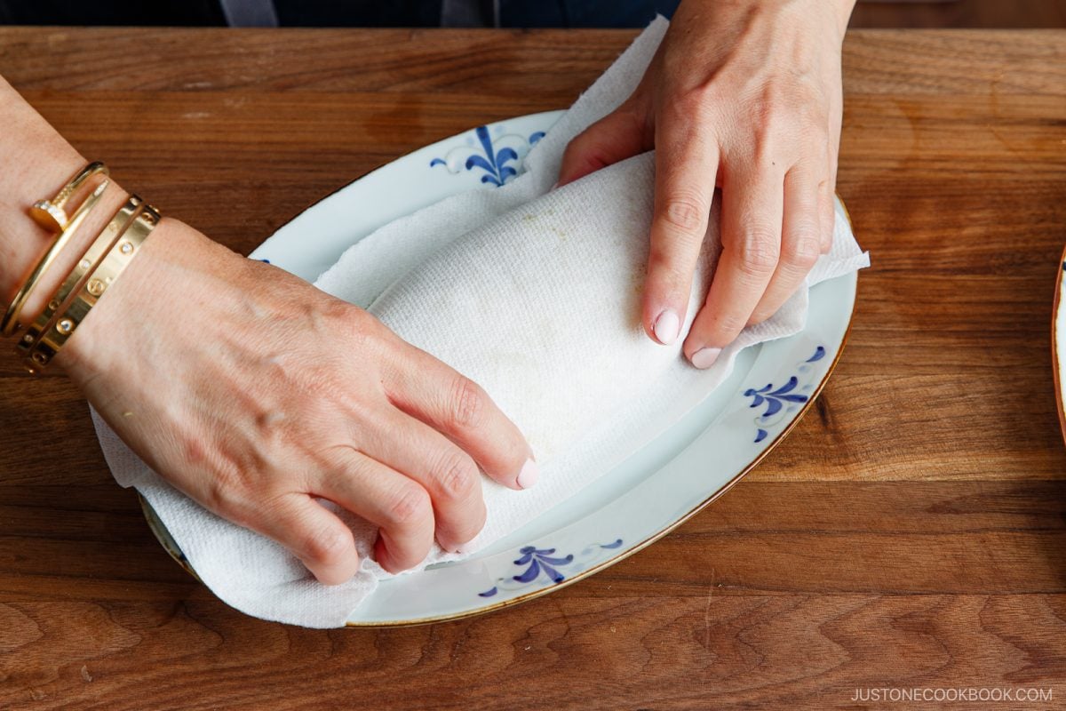 Two hands wrap a food item&mdash;likely a tonpeiyaki&mdash;in a white paper towel on a decorative oval plate placed on a wooden surface. The person is wearing gold bracelets.