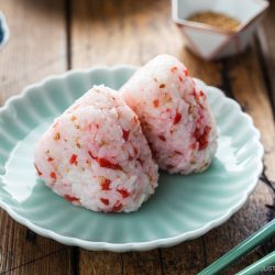 Two Umeboshi Onigiri (Plum Rice Balls), inspired by Midnight Diner, with red seasoning sit on a pale-green plate, accompanied by green chopsticks. A wooden table, pickled garnish, water glass, and small dishes are visible in the background.