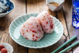 Two Umeboshi Onigiri (Plum Rice Balls), inspired by Midnight Diner, with red seasoning sit on a pale-green plate, accompanied by green chopsticks. A wooden table, pickled garnish, water glass, and small dishes are visible in the background.