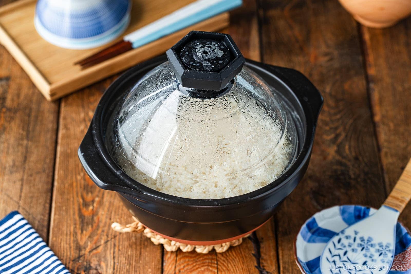 A black donabe clay pot with a glass lid filled with cooked white rice sits on a wooden table.