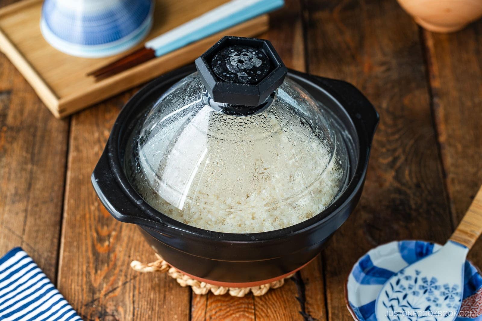 A black clay pot with a clear, condensation-covered glass lid sitting on a wooden table, containing cooked white rice.