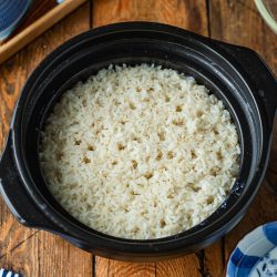 A black clay pot filled with cooked white rice sitting on a wooden table.
