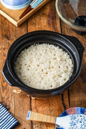 A black donabe filled with cooked white rice sits on a wooden table, next to a rice paddle and patterned dishware.