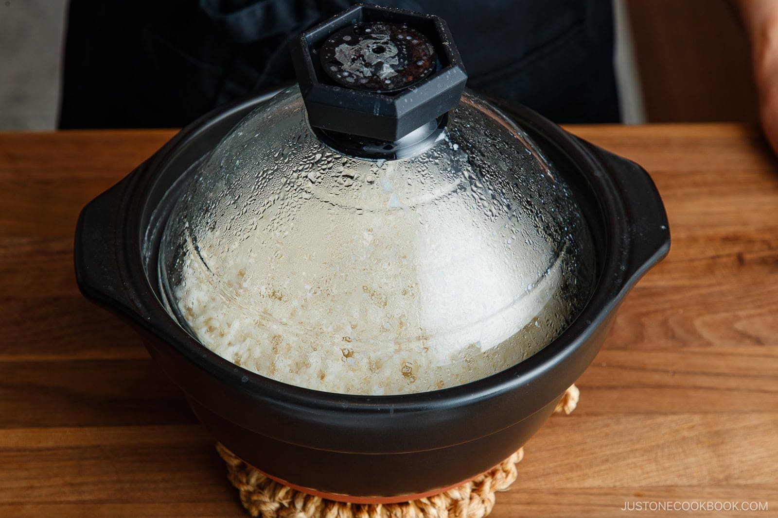 A black ceramic pot with a glass lid, containing steaming brown rice, sitting on a wooden surface.
