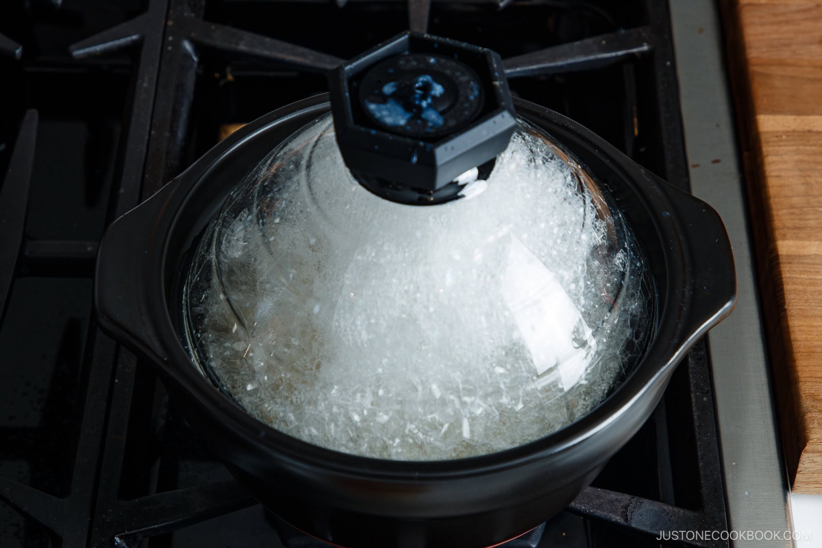 A black clay pot with a glass lid cooking rice on a stovetop.