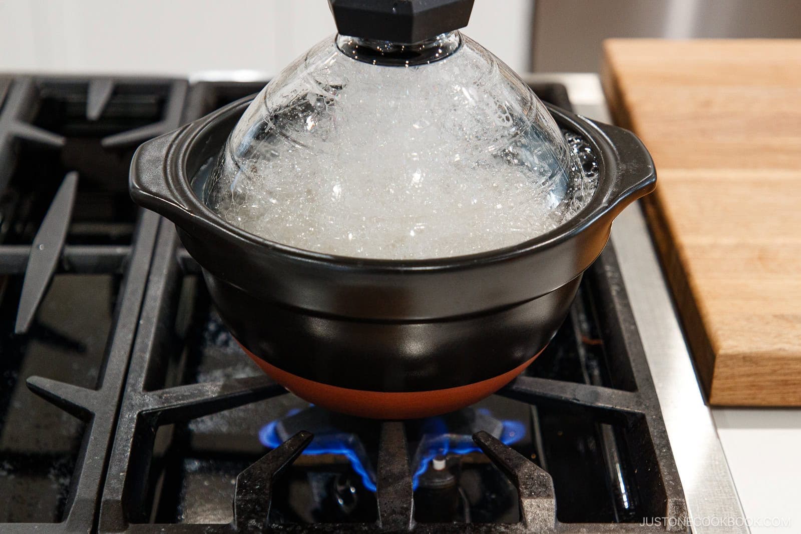 A round, black ceramic pot with a glass lid, filled with bubbling liquid, sits on a gas stovetop.