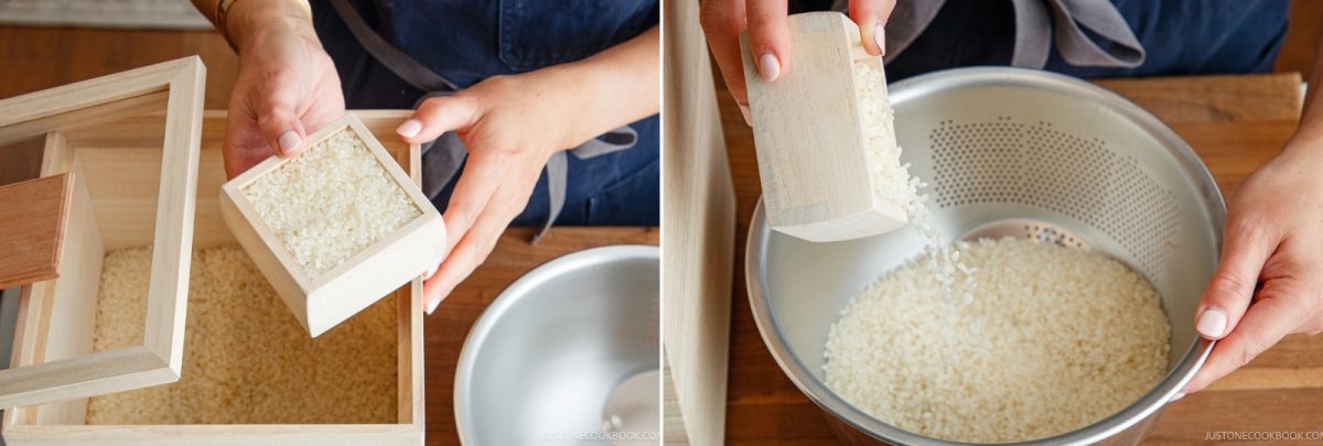 A person uses a wooden measuring box to scoop raw white rice from a wooden container, preparing to cook rice in a donabe by pouring the grains into a metal colander.