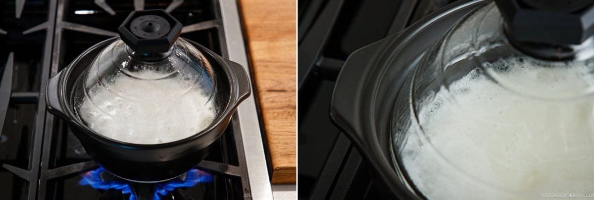 A black clay donabe with a clear glass lid containing boiling rice on a gas stove.