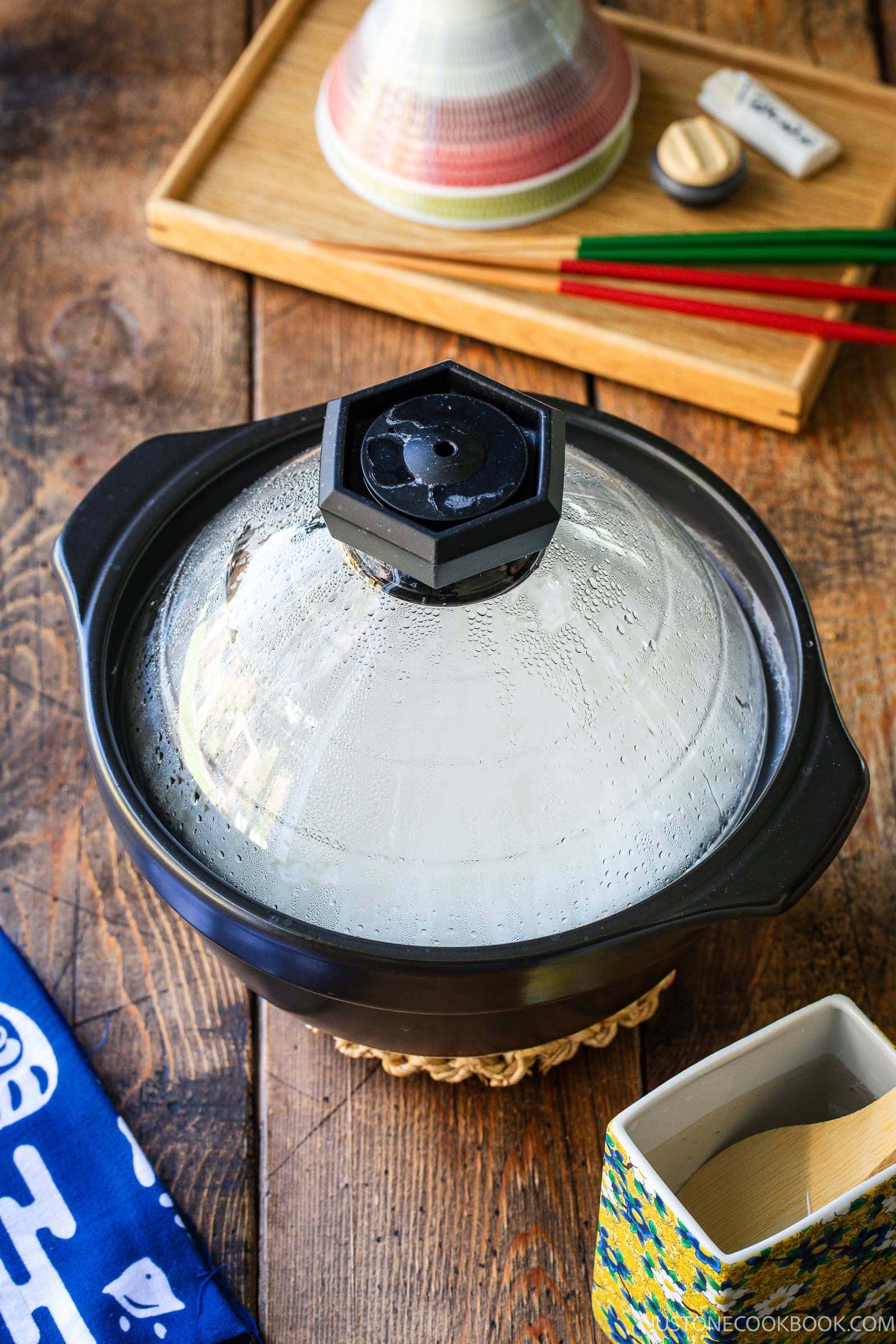 A black clay pot with a steaming glass lid sits on a wooden table next to a patterned container with a wooden spoon and a tray holding chopsticks.
