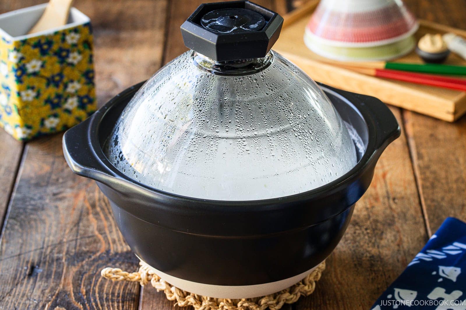 A black donabe clay pot with a domed glass lid sitting on a woven trivet on a wooden table.