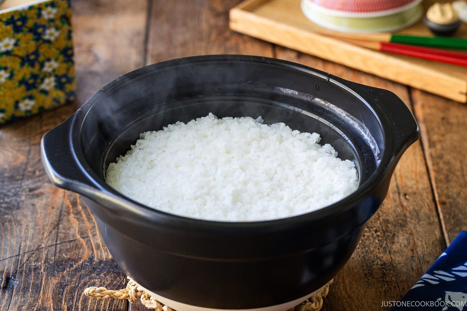 A steaming pot of freshly cooked white rice in a black ceramic donabe on a wooden table.