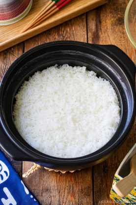 A black ceramic pot filled with cooked white rice sitting on a wooden table.