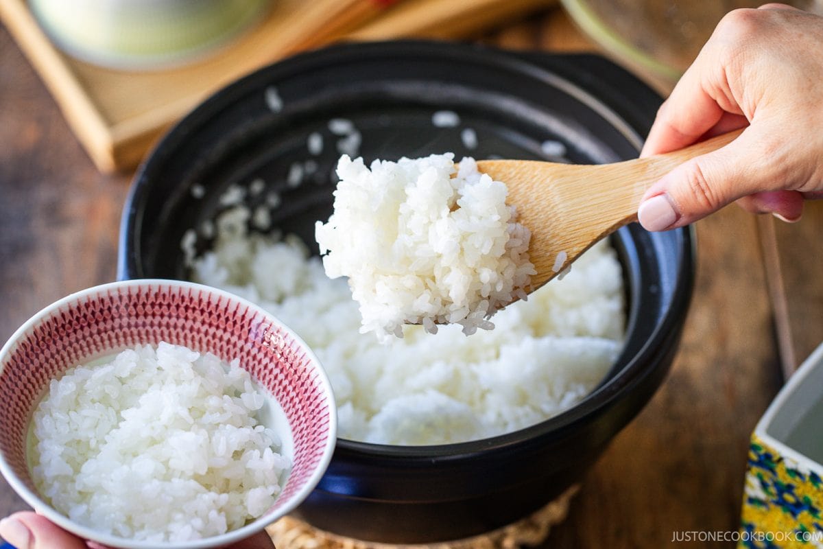 A hand uses a wooden spoon to serve steamed white rice from a black clay pot into a small, patterned bowl.