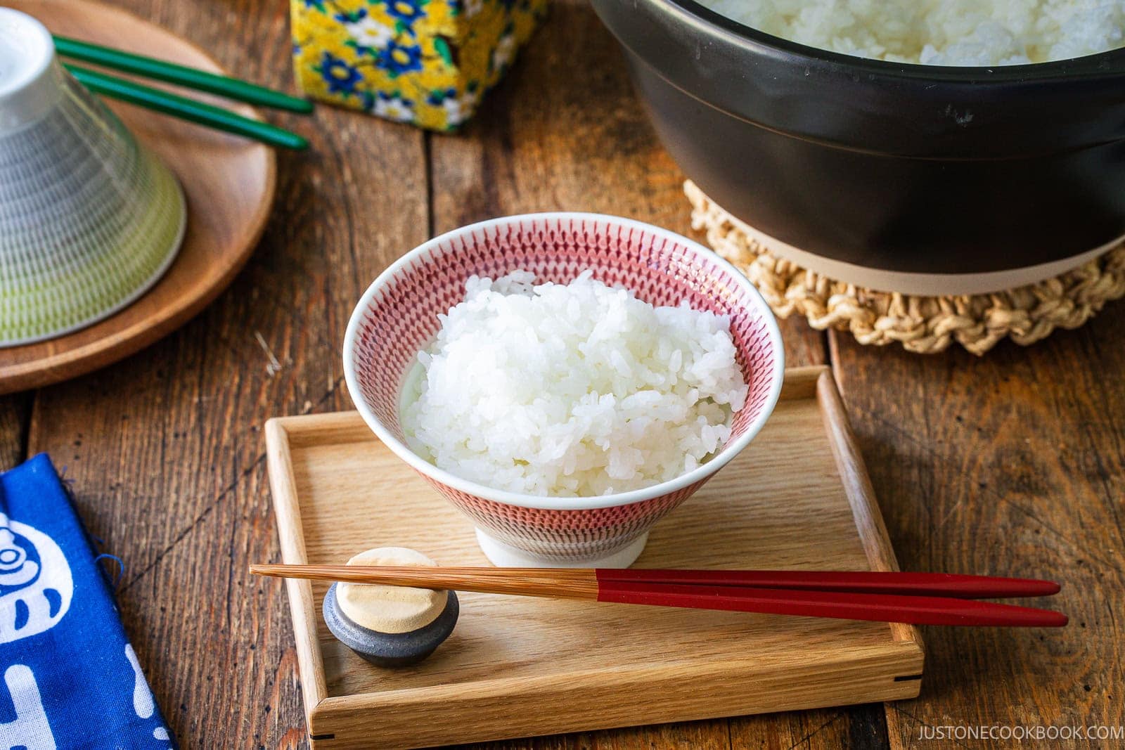 A small bowl of cooked white rice sits on a wooden tray with a pair of red chopsticks resting on a ceramic holder.