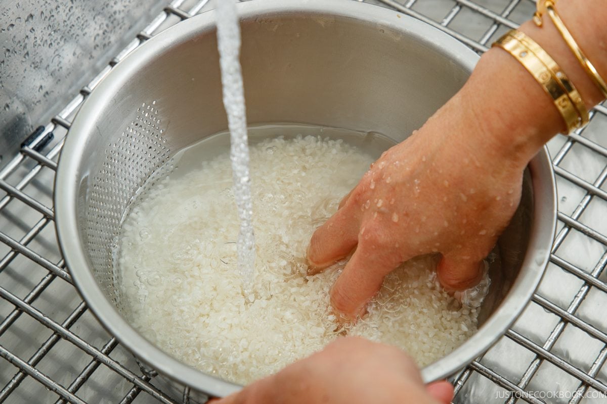 A hand rinses white rice in a metal colander under a running kitchen faucet as the first step to cook rice in a donabe.
