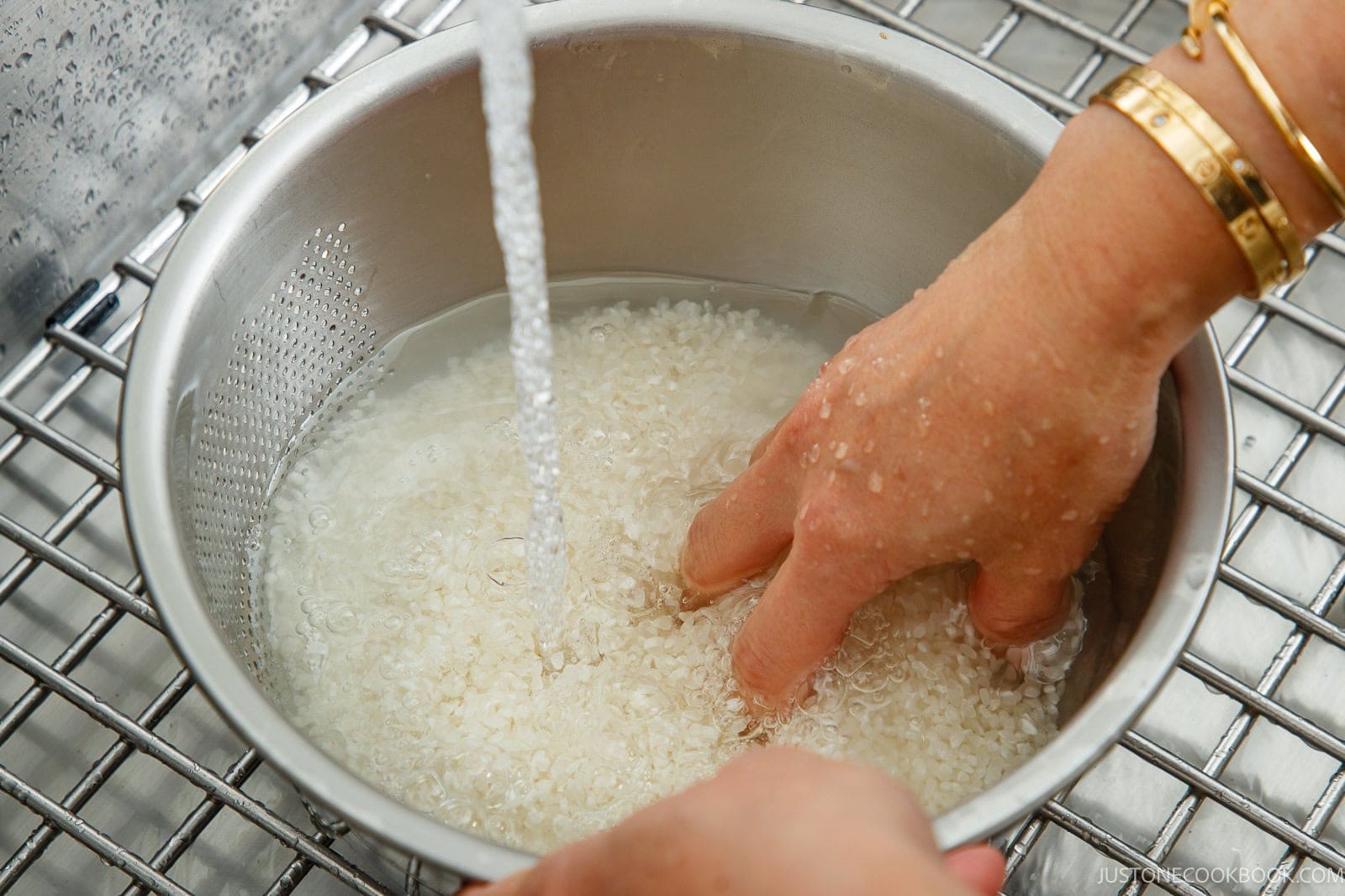 A hand rinses white rice in a metal colander under a running kitchen faucet as the first step to cook rice in a donabe.