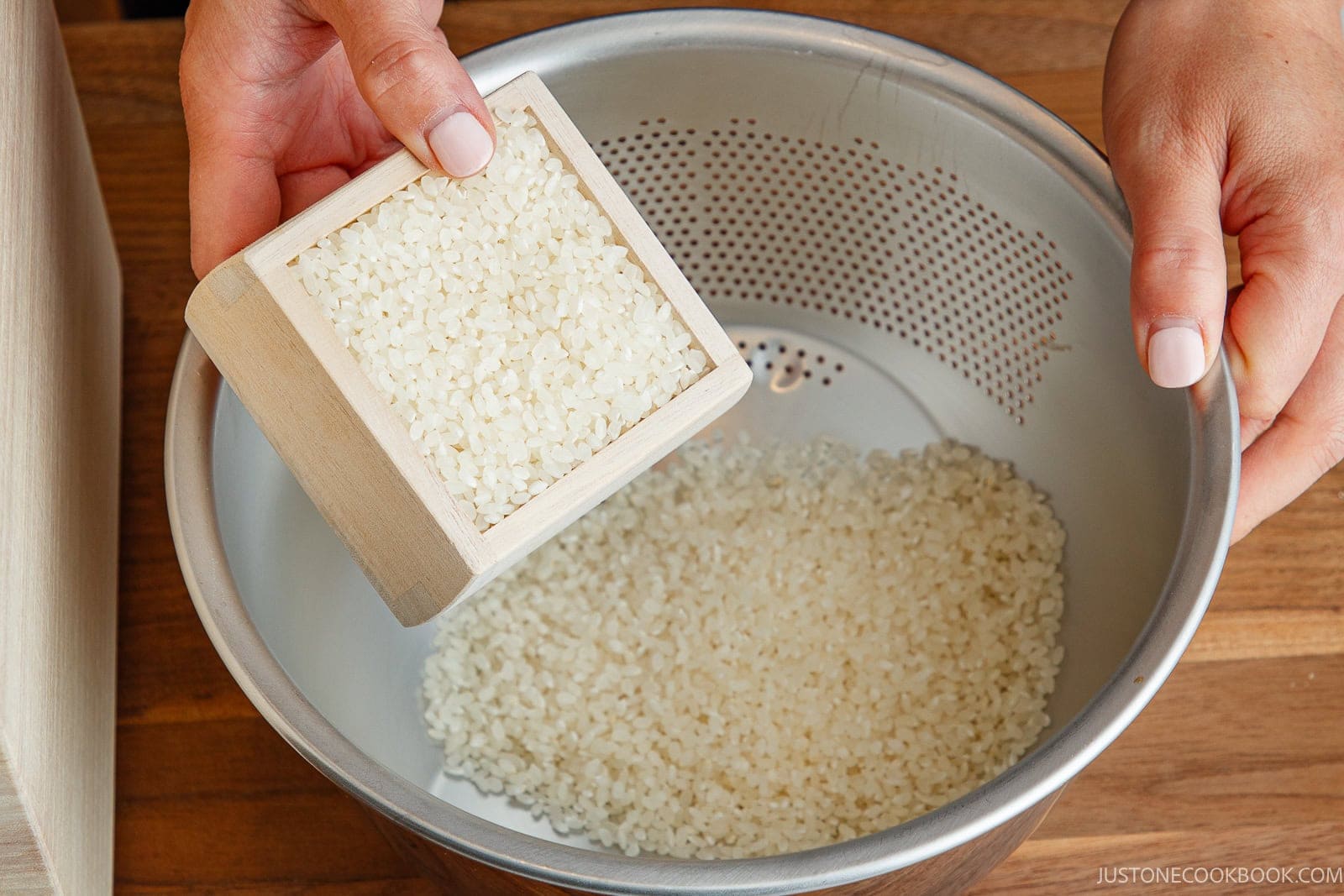 Hands pouring white rice from a wooden measuring box into a metal colander to prepare it for how to cook rice in a donabe.