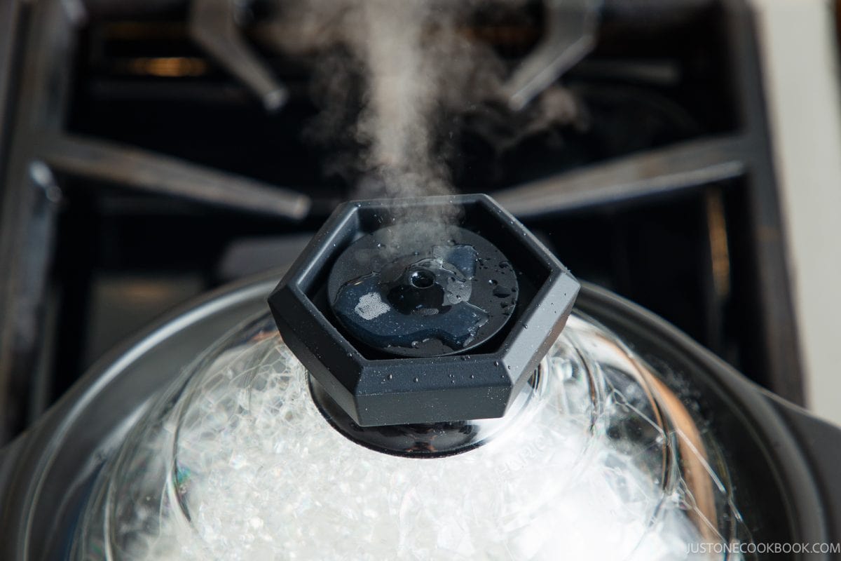 Steam escapes from the vent of a clear glass pot lid as you learn how to cook rice in a donabe on the stovetop.