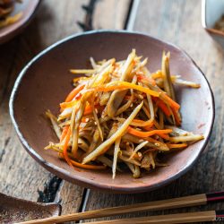 A bowl of Kinpira Gobo (Braised Burdock Root), a classic Japanese dish featuring saut&eacute;ed burdock root and carrots, sits on a wooden table with chopsticks beside it.