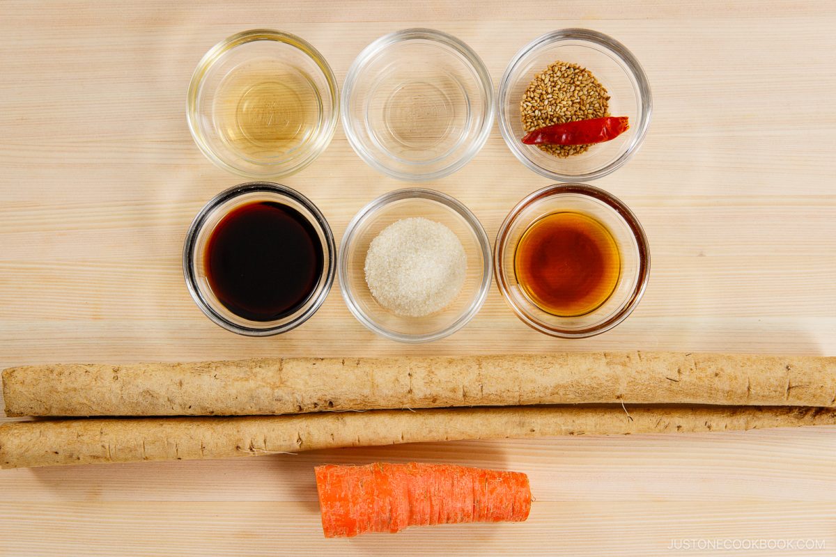 Six small bowls with sauces, vinegar, sugar, and sesame seeds are arranged above two long burdock roots and a piece of carrot on a light wooden surface&mdash;perfect ingredients for preparing Kinpira Gobo (Braised Burdock Root).