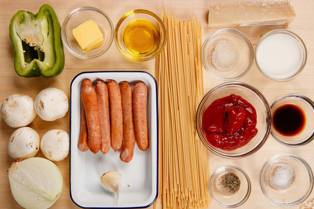 Ingredients for a pasta dish arranged on a wooden surface, including sausages, spaghetti, green bell pepper, mushrooms, onion, butter, oil, parmesan, milk, ketchup, soy sauce, garlic, and seasonings.