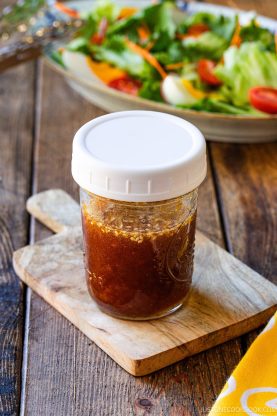 A mason jar filled with brown wafu dressing sits on a small wooden cutting board. In the blurred background, a fresh salad with greens, tomatoes, and carrot strips is visible on a plate.