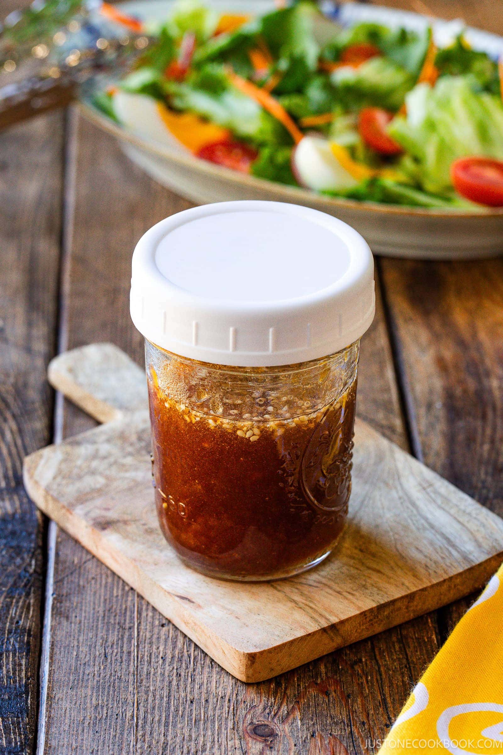 A mason jar filled with brown wafu dressing sits on a small wooden cutting board. In the blurred background, a fresh salad with greens, tomatoes, and carrot strips is visible on a plate.