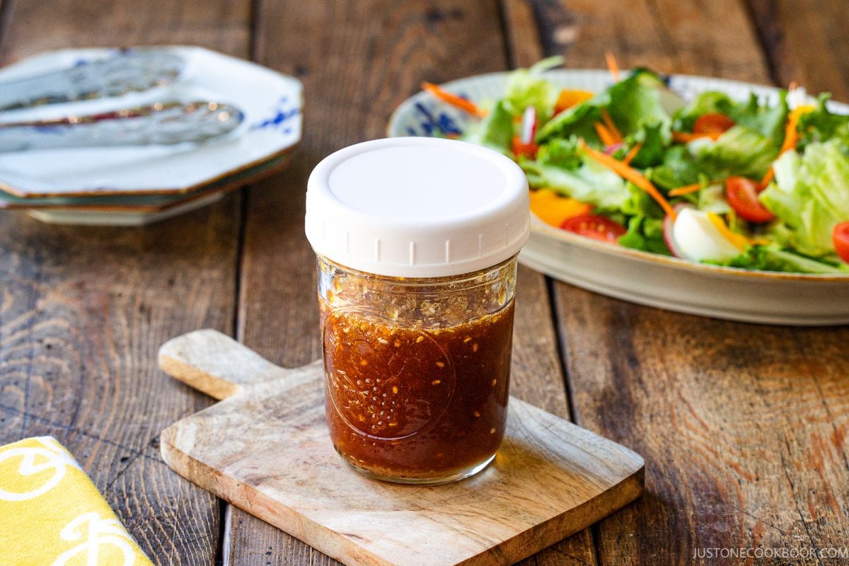 A jar of homemade wafu dressing sits on a small wooden board, with a fresh salad and plates in the background on a rustic wooden table.