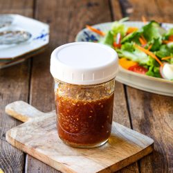 A jar of homemade wafu dressing sits on a small wooden board, with a fresh salad and plates in the background on a rustic wooden table.