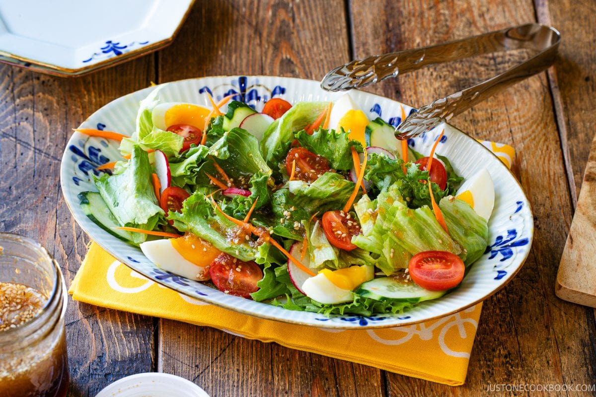 A colorful salad with leafy greens, cherry tomatoes, sliced cucumbers, carrots, and boiled egg pieces on a patterned plate is served with wafu dressing, with salad tongs on a rustic wooden table.
