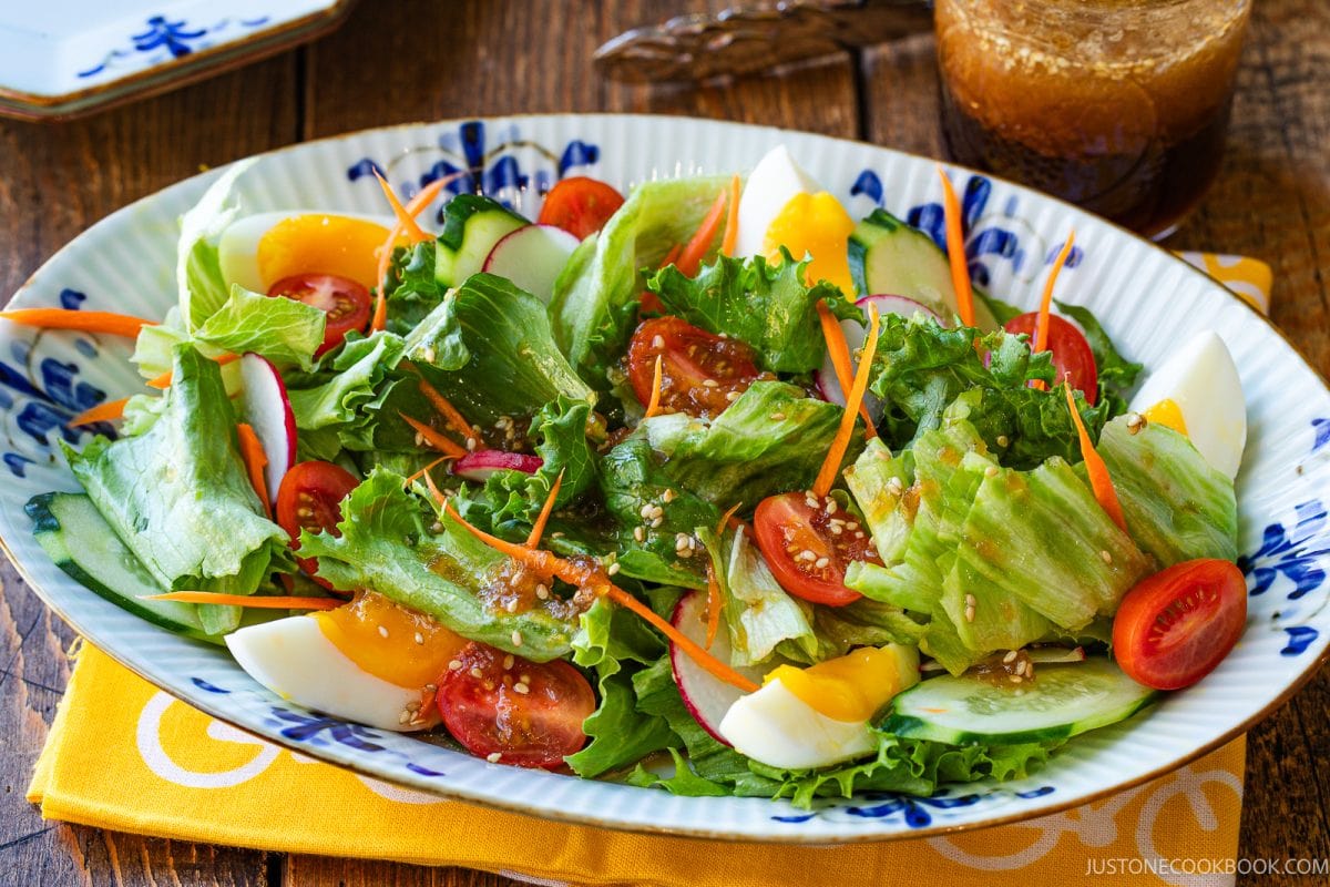 A salad with lettuce, cherry tomatoes, radish, cucumber, carrots, and boiled egg slices is served on an oval plate, accompanied by wafu dressing in the background.