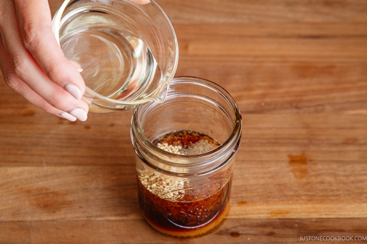 A hand pours oil from a glass bowl into a mason jar containing a dark sauce with sesame seeds on a wooden surface.